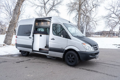 RV rental: Silver Mercedes Sprinter camper van with open side door showing interior, parked in winter snow.
