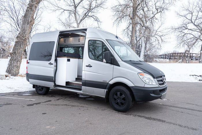 RV rental: Silver Mercedes Sprinter camper van with open side door showing interior, parked in winter snow.