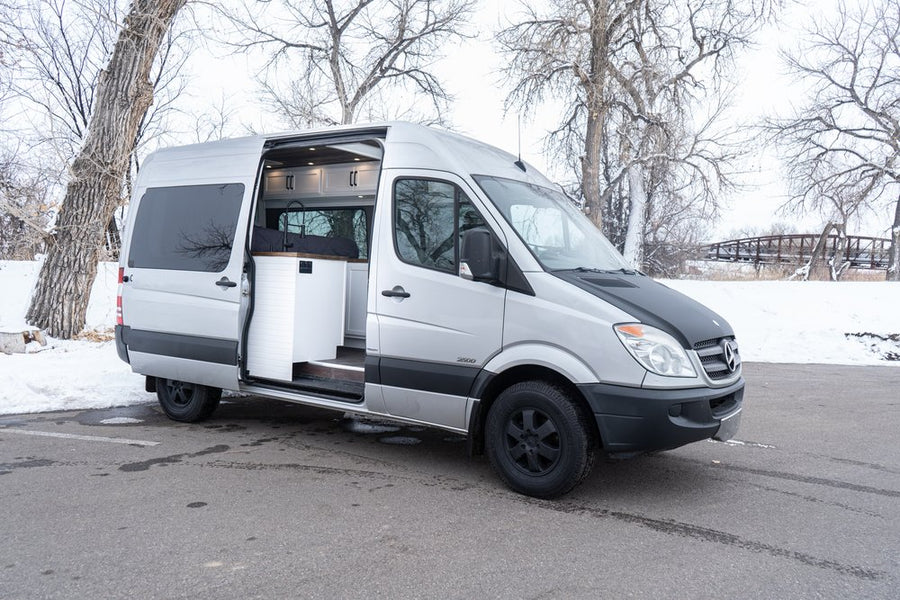RV rental: Silver Mercedes Sprinter camper van with open side door showing interior, parked in winter snow.