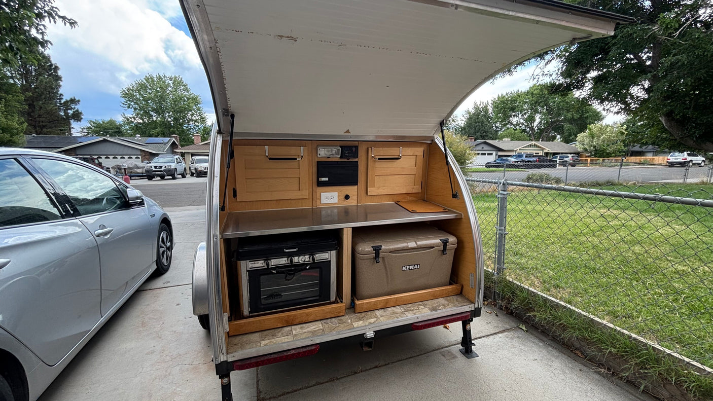 Teardrop trailer's open galley kitchen shows an oven, stove, and cooler, offering compact outdoor cooking for RV rental.
