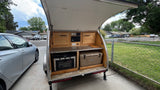 Teardrop trailer's open galley kitchen shows an oven, stove, and cooler, offering compact outdoor cooking for RV rental.