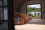 Enjoy this cozy rv rental bed setup with pillows & a blanket, overlooking a scenic lake with a campfire, chairs, and string lights.