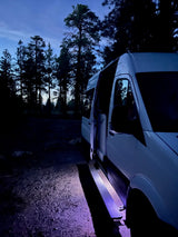 White **RV rental** van parked at night with open door and glowing purple step in a dim forest. Ready for adventure.