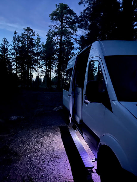 White **RV rental** van parked at night with open door and glowing purple step in a dim forest. Ready for adventure.