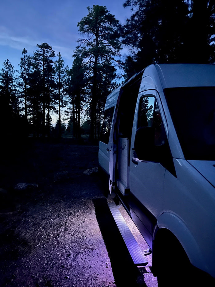 White **RV rental** van parked at night with open door and glowing purple step in a dim forest. Ready for adventure.