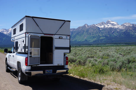 Wyoming's Grand Tetons frame this white truck camper RV rental on a dirt road. Perfect for mountain adventures.