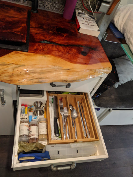 Compact RV rental living: Custom wood counter and open drawer revealing organized cutlery, spices, and utility items.