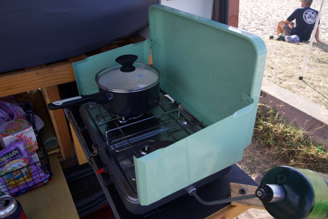 Preparing a meal on a portable camping stove, likely from an RV rental. Snacks nearby; a person relaxes in the background.