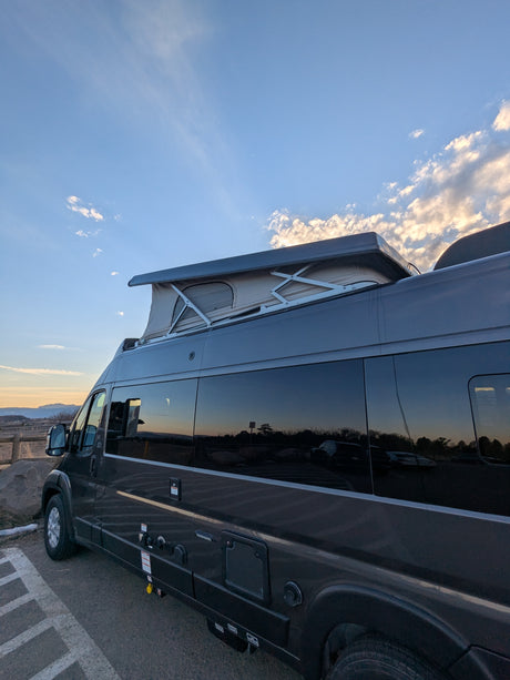 Dark grey RV rental with an extended pop-top tent, parked on asphalt. Sunset sky reflects in windows.