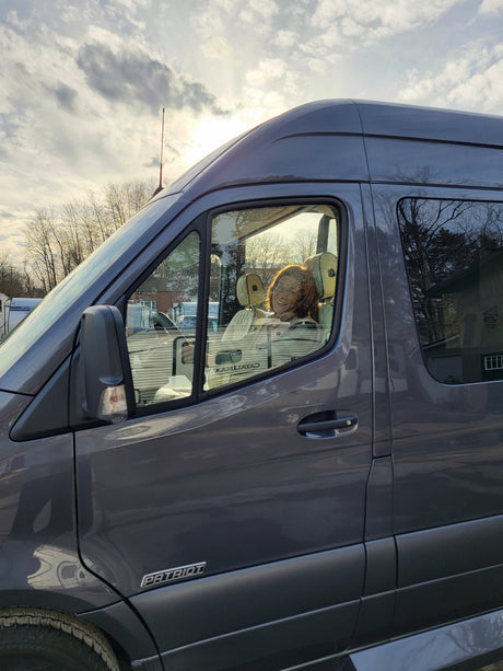 A grey RV rental camper van parked outdoors, showing a driver's side view with bright sun reflecting off the windows, a person visible inside.