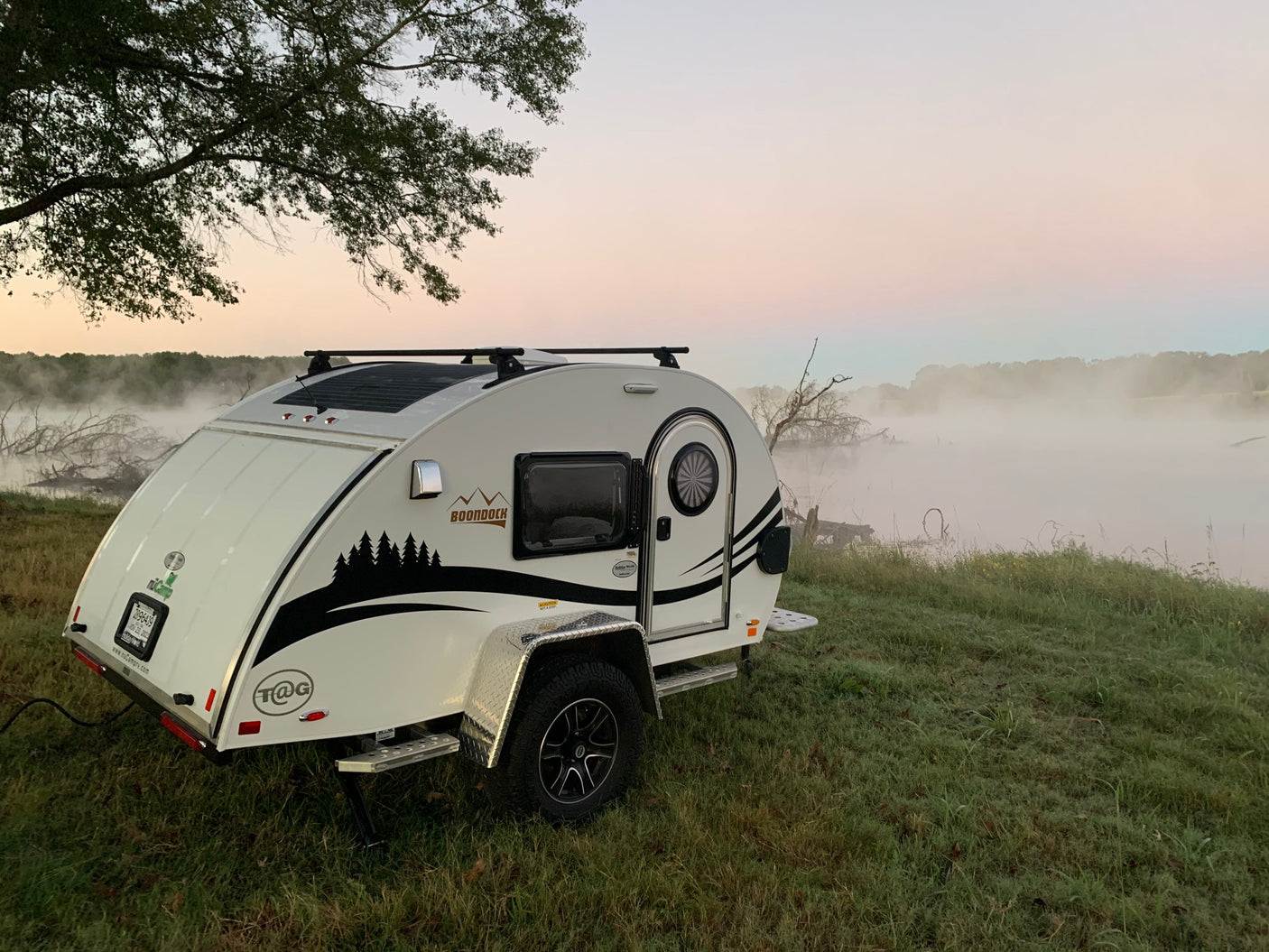 Misty sunrise scene: a white teardrop RV rental parked by foggy water, highlighting serene camping.