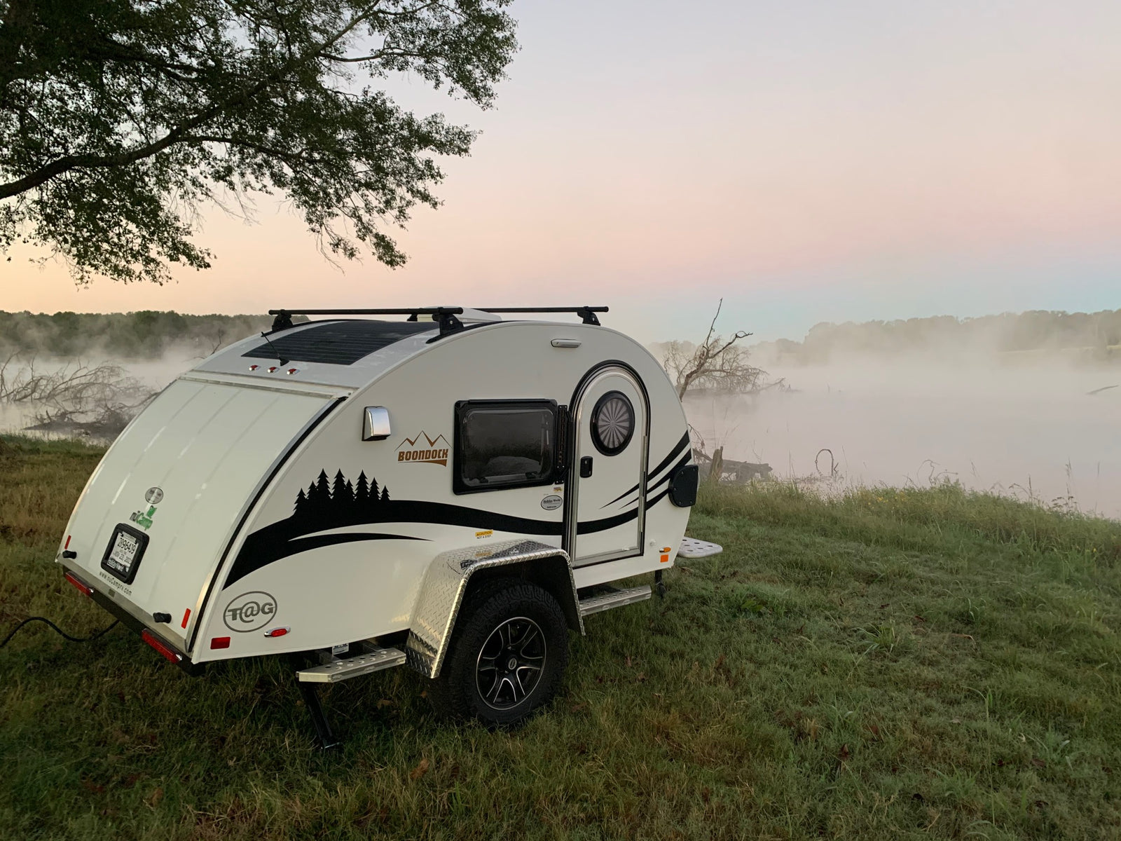 Misty sunrise scene: a white teardrop RV rental parked by foggy water, highlighting serene camping.