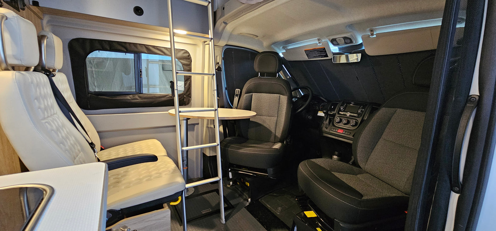 Spacious rv rental interior: view showing driver's cabin with dark seats, and light living area seats, table, and bunk ladder.