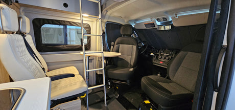 Spacious rv rental interior: view showing driver's cabin with dark seats, and light living area seats, table, and bunk ladder.