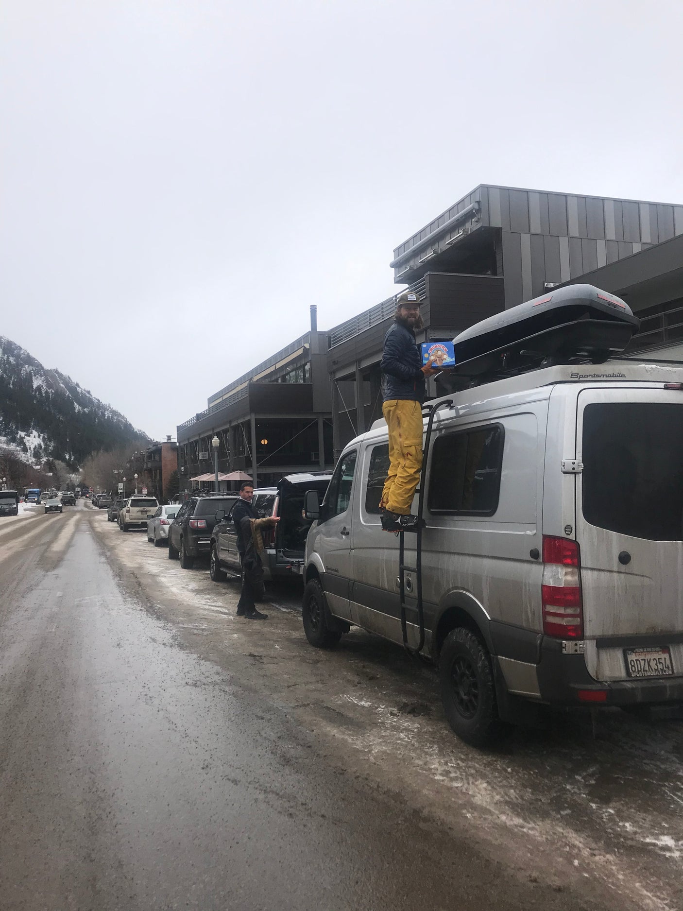 Man on an RV rental's ladder holds Drumsticks on a slushy street in a snowy mountain town.