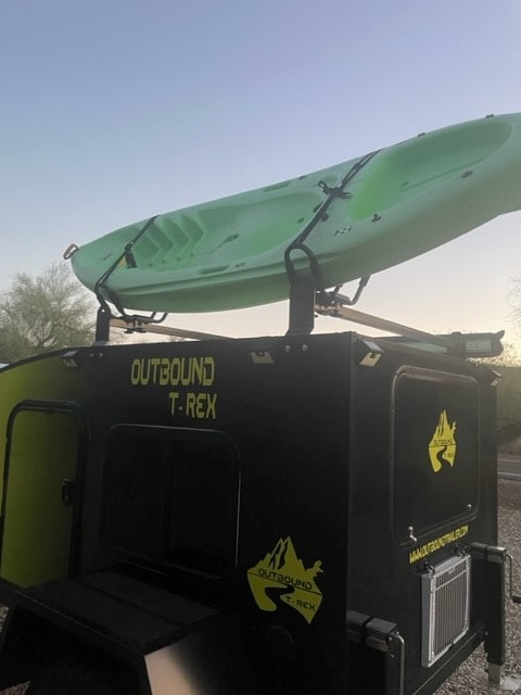  and camping gear on a rug.|Outbound T-Rex adventure trailer for rv rental with kayak on roof. Open doors reveal sleeping area and rear storage.|Bright yellow Outbound T-Rex teardrop trailer RV rental with outdoor kitchen — photo 7