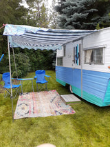 This inviting rv rental offers a comfortable outdoor setup with a striped awning, colorful rug, and blue chairs on green grass.