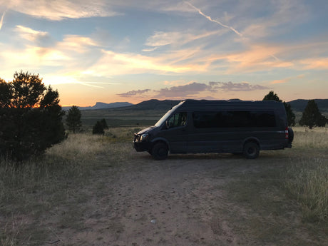 Experience freedom with an RV rental! A dark van is parked on a dirt path, capturing a serene sunset over mountains.