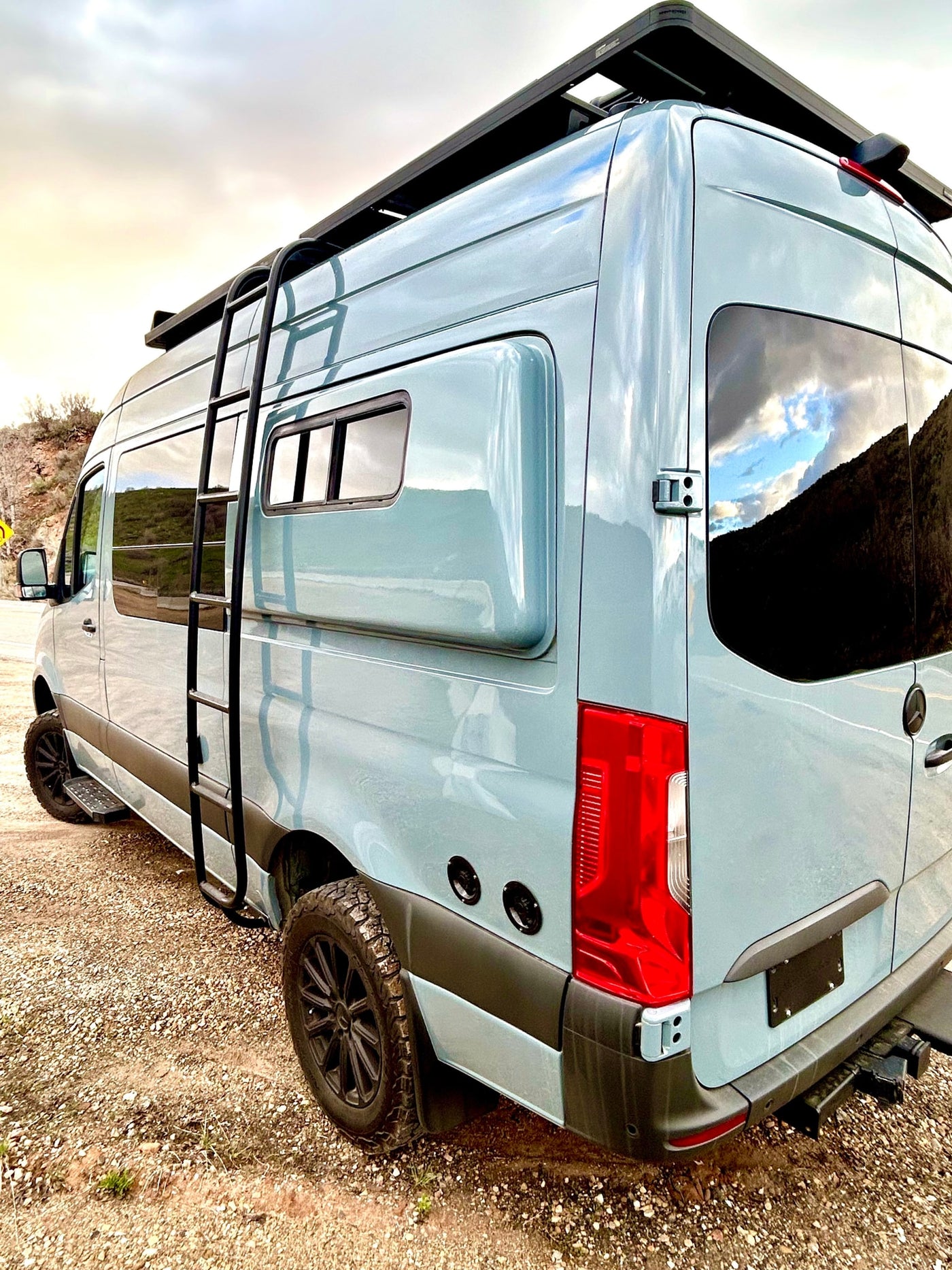 This adventure-ready camper van, perfect for rv rental, features a ladder, roof rack, and off-road tires, parked on gravel. — photo 10