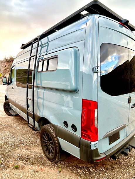 This adventure-ready camper van, perfect for rv rental, features a ladder, roof rack, and off-road tires, parked on gravel.