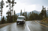 White camper van (RV rental) on a wet, winding mountain road. Tall pines and distant peaks create a scenic travel vista.