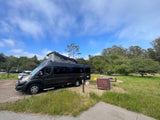 Dark grey camper van RV rental with pop-top roof extended at a sunny campsite amidst green grass & trees.
