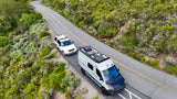 Modern Sprinter van rv rental, equipped with solar panels, follows a white SUV on a winding, nature-filled road.
