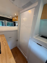 Explore this RV rental's modern interior: raised bed, wood kitchen counter, and sleek white bathroom/shower door.