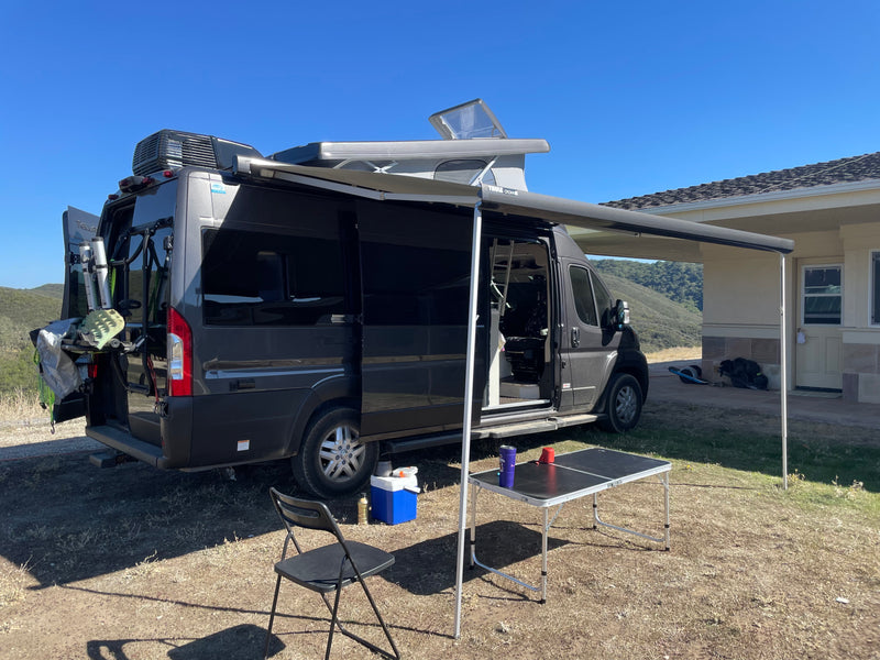 RV rental: Dark gray camper van with pop-top roof, extended awning, and outdoor camp setup (table, chair, cooler). Gear on back. — photo 2
