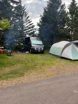 Enjoying a serene campsite with an RV rental, a tent, and a warm campfire. Smoke rises among the tall pines.
