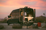 Cozy RV rental camper van at sunset, ready for adventure! Back doors open to a bed and camping gear in a desert landscape.