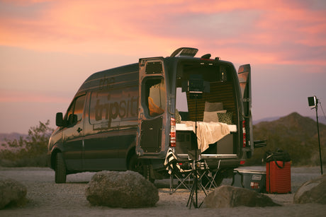 Cozy RV rental camper van at sunset, ready for adventure! Back doors open to a bed and camping gear in a desert landscape.