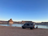 Scenic RV rental adventure! Gray camper van with bikes by a lake, surrounded by stunning rock formations under a blue sky.