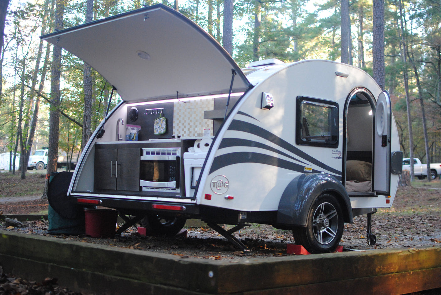 This compact teardrop camper, ideal for an RV rental, displays its open rear kitchen and open side door in a wooded campground.