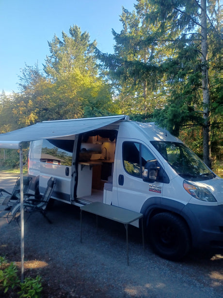 RV rental van, parked under trees with an extended awning and outdoor setup. Its open door reveals a functional kitchen, ready for van life.