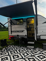 Evening at a Catalina RV rental: Awning with blue lights, steps, grill, chair, and patterned outdoor mat.