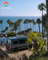 An RV rental Moterra campervan is parked on a dirt lot, offering a scenic view of the palm-lined ocean.
