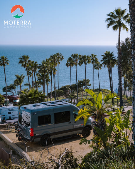 An RV rental Moterra campervan is parked on a dirt lot, offering a scenic view of the palm-lined ocean.