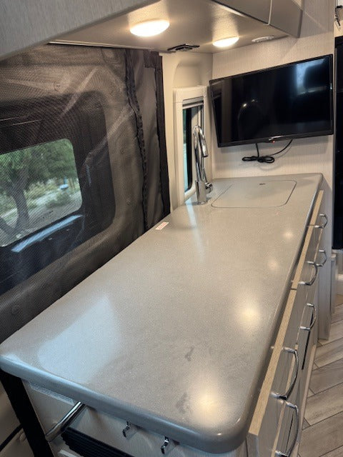 Modern rv rental kitchen with gray countertop, integrated sink, mounted TV, and dark mesh window screen on the left.