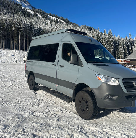 A sleek gray camper van, an excellent RV rental for snowy mountain getaways, parked amidst snow-covered pines under a clear blue sky.