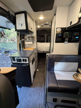 RV rental camper van interior featuring a kitchenette, dinette, and bed, with a scenic river view outside.