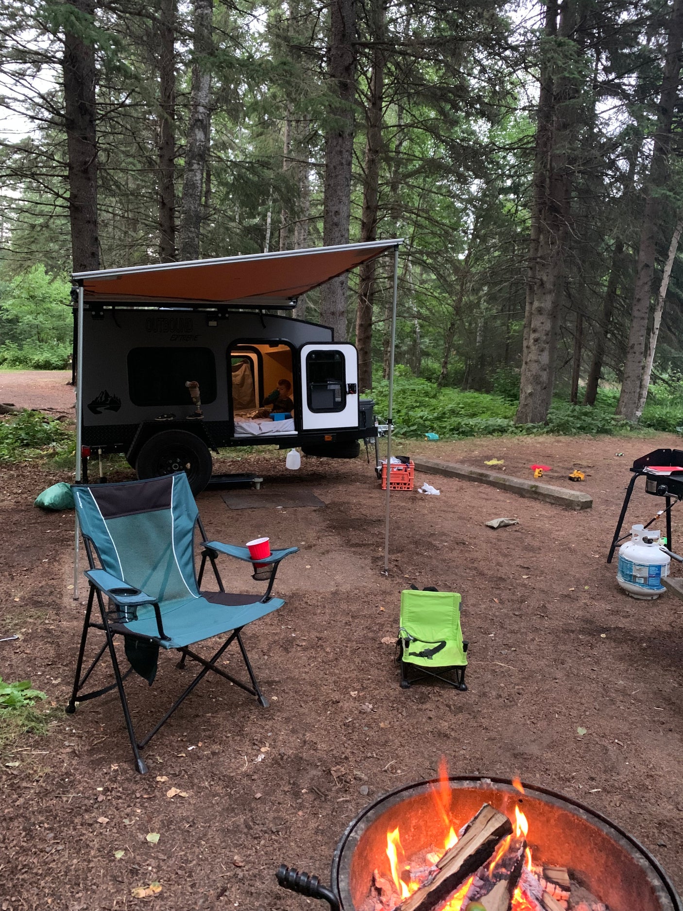 Forest campsite scene with an RV rental, featuring a child inside the camper, a glowing campfire, and camping chairs.