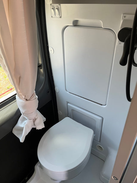 An RV rental features a compact wet bath with toilet, shower head, and light/fan switches. A window offers natural light.