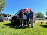 A family smiles with their red RV rental van on a sunny day at a grassy campground with other RVs nearby.