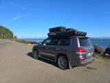 Explore with an RV rental: Dark gray overlanding SUV parked roadside, featuring roof-top cases & awning by a scenic bridge.
