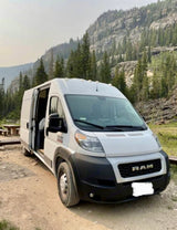 Explore nature with this Ram ProMaster RV rental. White van parked on a dirt road, surrounded by scenic mountains.