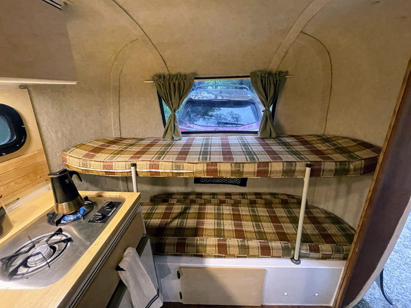 Inside a compact RV rental, featuring a functional kitchen with a stove and cozy bunk beds with plaid cushions.