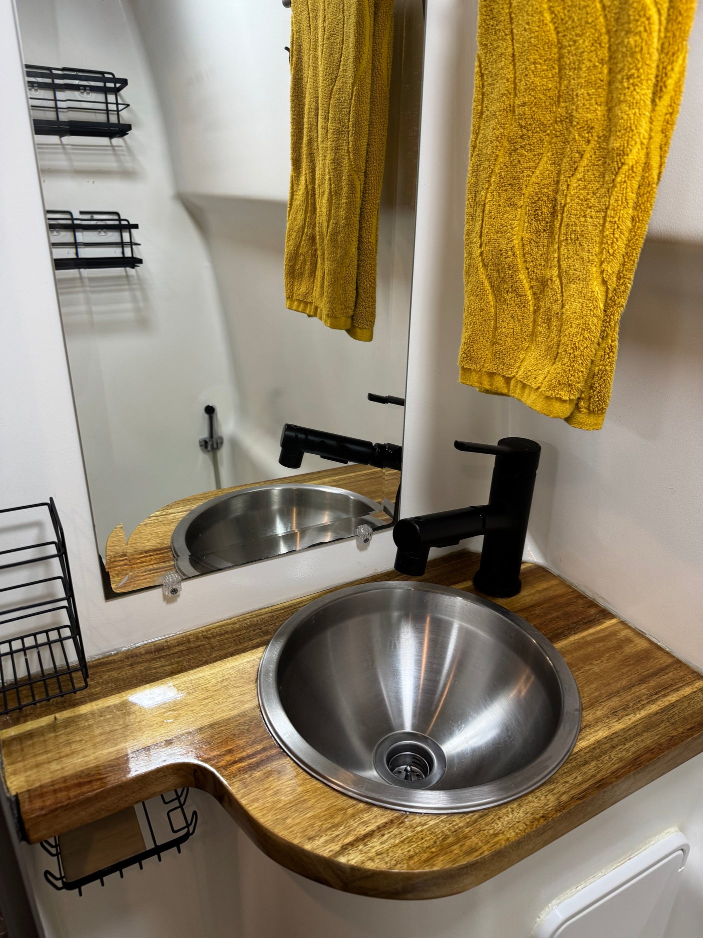 A modern, compact RV rental bathroom with a stainless steel sink, black faucet, and wooden counter. Yellow towels hang beside the mirror.