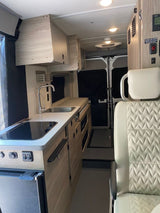 Explore this modern RV rental interior! Features a compact galley kitchen, overhead storage, and passenger seat, perfect for road trips.