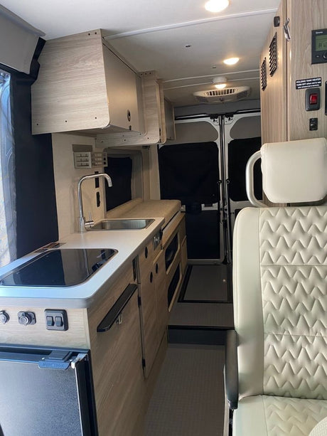 Explore this modern RV rental interior! Features a compact galley kitchen, overhead storage, and passenger seat, perfect for road trips.